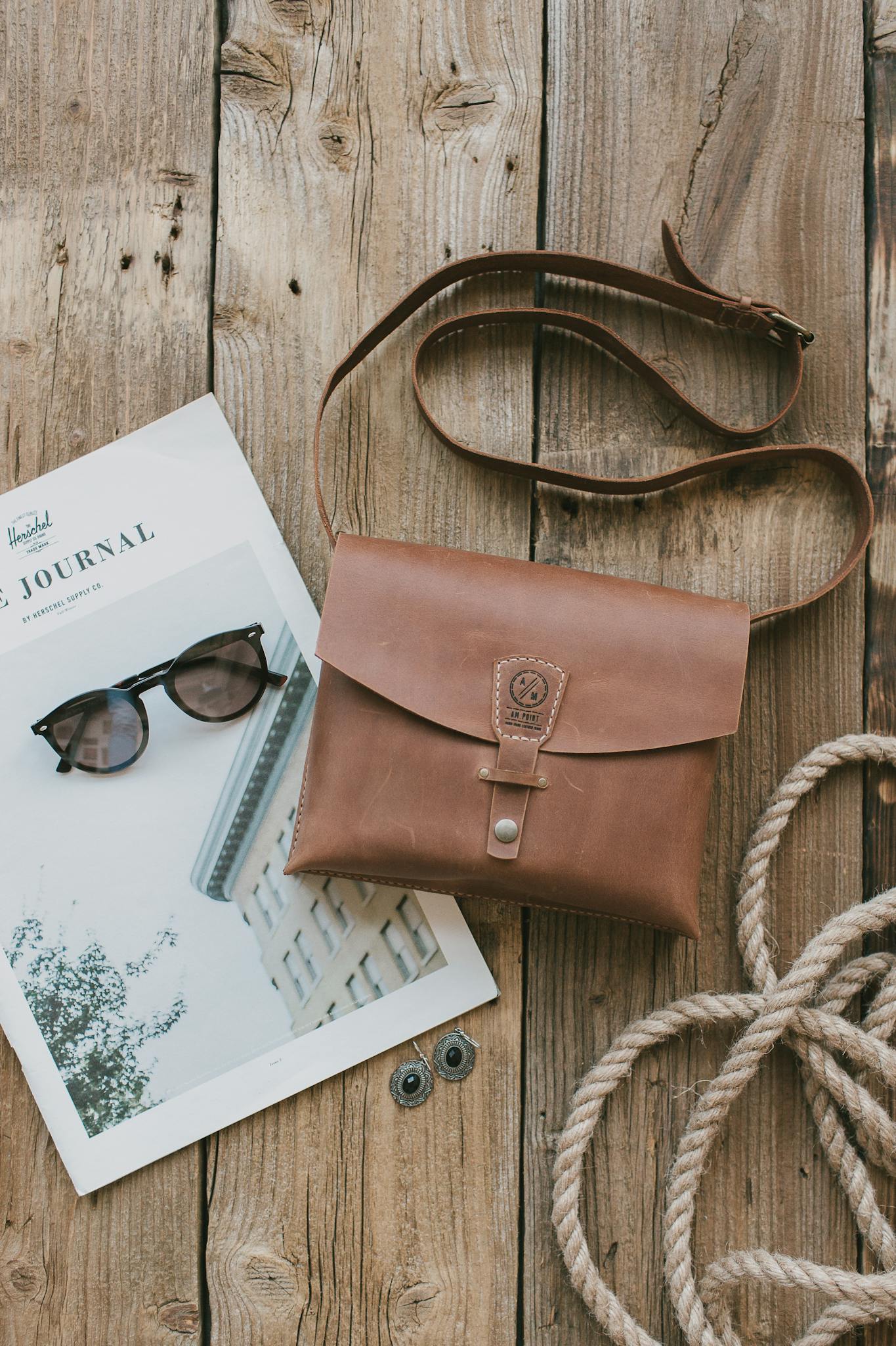 A stylish flat lay featuring a leather sling bag, rope, sunglasses, and a magazine on a rustic wooden table.